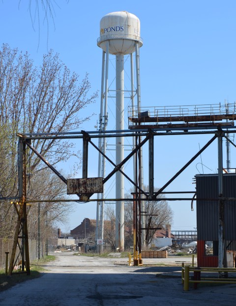 white water tower in the background with Ponds written on it, metal overhead structure for trucks entering old abandoned factory in the foreground with faded sign that once was warning speed bumps 