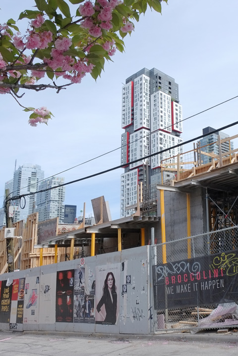 construction site, with posters on the hoardings, tall white, grey, and red building in the background. 