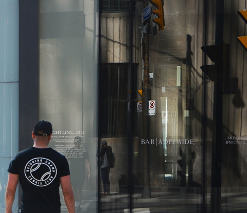 a man in a black tshirt crosses the street towards a large indow with lots of reflections in it.