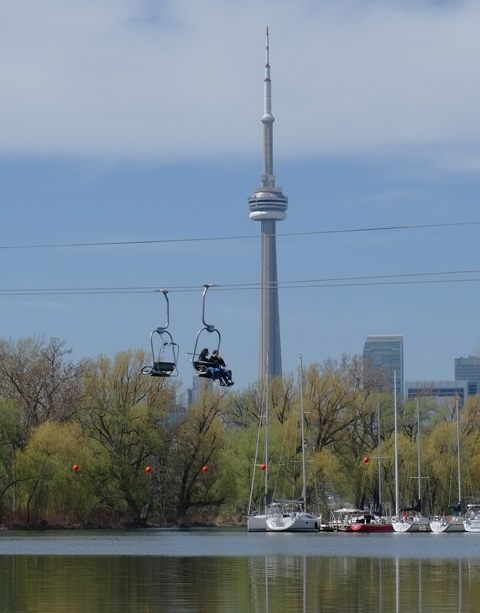 CN tower in the background, people on the Skyline ride at Centre Island passing over water, with large boats docked farther up the river 