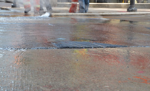 wet road, water gushing out of a large hose, feet and legs of some men.