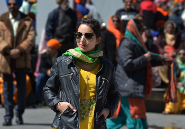 a young sikh woman stands in front of a crowd of people, yellow sari, black leather jacket and green and gold scarf around her neck 