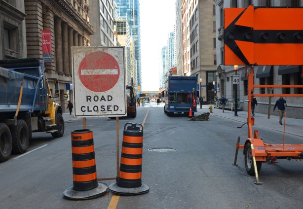 road closed sign, black arrow on orange sign, ornage and black striped traffic cones, blocking Yonge street, with trucks in the background. 