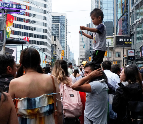 a boy on his father's shoulders, playing with a green bead necklace as they watch the pride parade in TOronto 