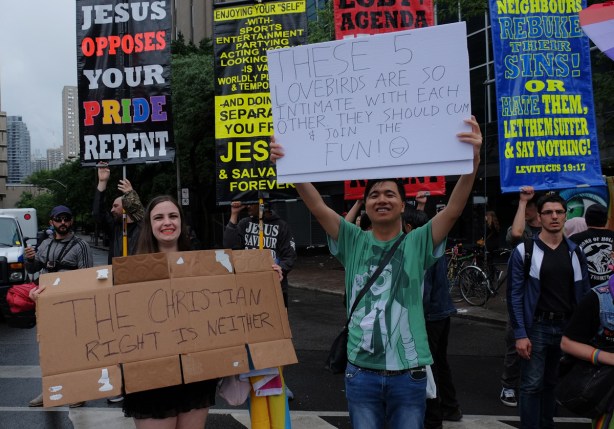 two protestors hold up hand written signs to block the protest behind them. One sign, on brown cardboard, says The Christian right is neither. THe other sign, on white bristol board says These 5 lovebirds are so intimate with each other they should cum and join the fun.  One of the anti-pride posters says Jesus opposes your pride. repent.  