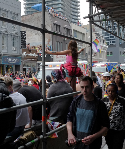 people watching pride parade on yonge street in toronto, scaffolding for construction, people on roofs, people walking by on sidewalk 