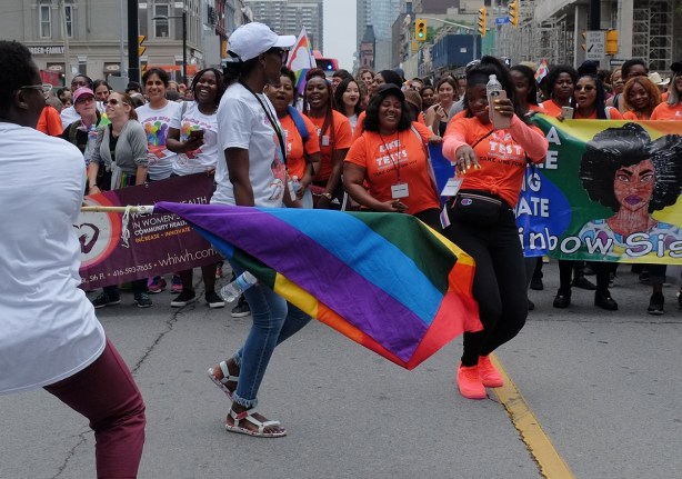 dyke march 2018 - dancing women in orange T-shirts, rainbow sisters, one is waving a rainbow flag 