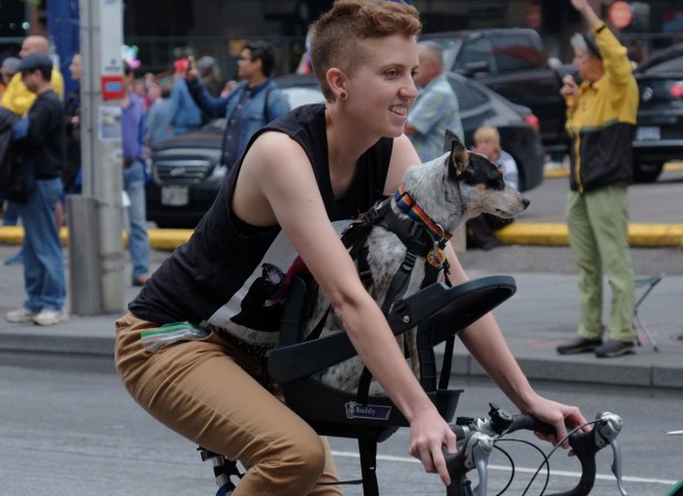 a woman with very short hair on bike with a dog sharing her seat, black and white dog with nose pointed forwards as they ride together 