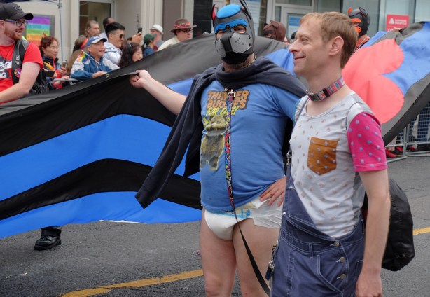 carrying a black and blue striped flag, two men, one in diaper and dog leather mask, the other in ordinary clothes 