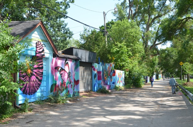 a lane with many painted garage doors, butterfly murals, summer time, green leaves, lots of trees, two people walking