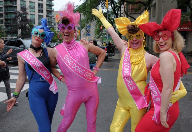 four people in stretchy tight fitting unitards, a pink, a blue, a yellow and a red, all have sashes that say Ding Dong Girls 