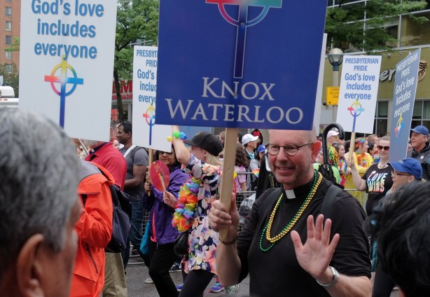marchers from the presbyterian church walking in the pride parade, man holding a sign that says Knox Waterloo, in clericals, with beaded necklaces around his neck 