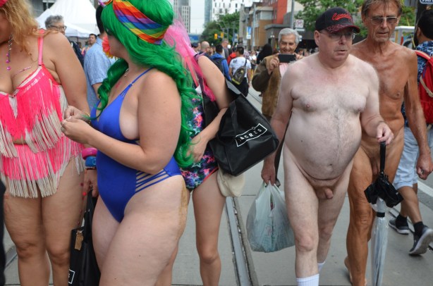 two naked men walking down the street, with a man behind them taking their picture, two women in bathing suits and large hats 