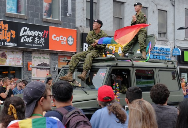 two military personnel sitting on top of a green jep with rainbow flags as they drive in the pride parade 