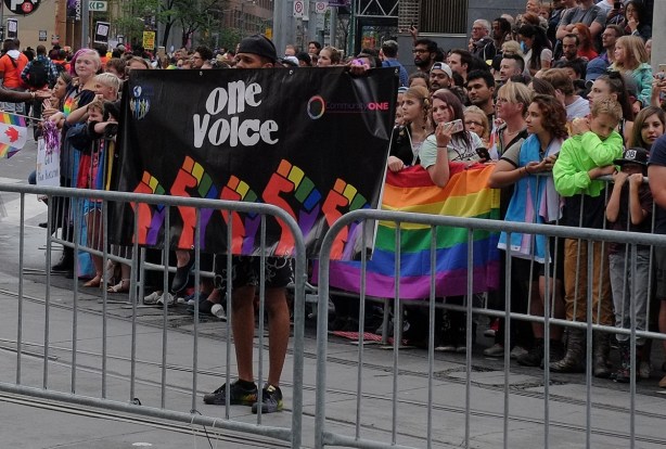 a young man in the pride parade stops to hold up a banner that says one voice. He is facing anti pride parade protestors 