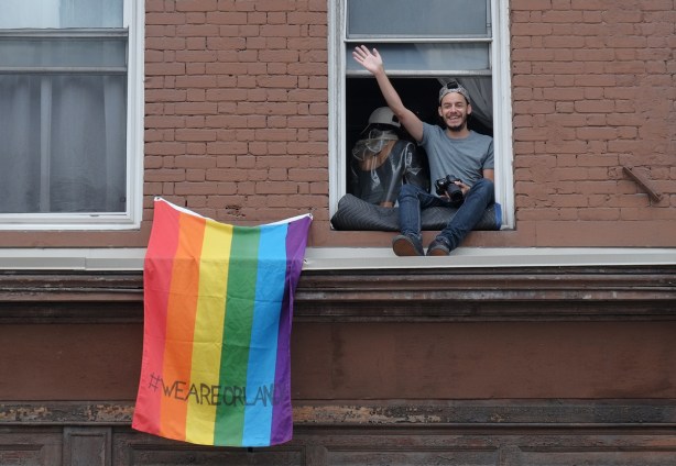 dyke march 2018 - a young man sits on the ledge of an open upper storey window on Yonge street, rainbow flag with #weareorlando (we are orlando) written on it 