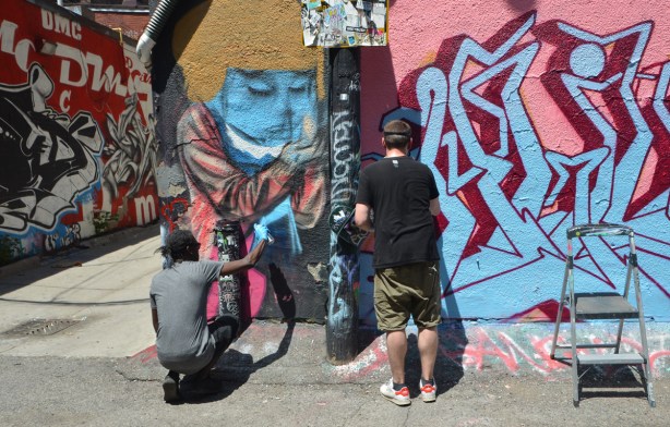 elicser paints a person on a wall, another man is beside him, also spray painting street art in Graffiti Alley 