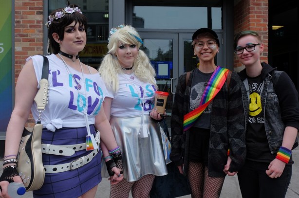 4 people on the sidewalk watching the dyke march Two of them are wearing white T shirts with L is for love on it in blue and purple letters, also flower garlands in their hair. 