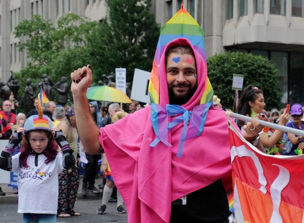 a man holds a banner with one arm, has the other arm lifted up, pink fabric draped around his shoulders, and a rainbow flag around the back and top of his head 