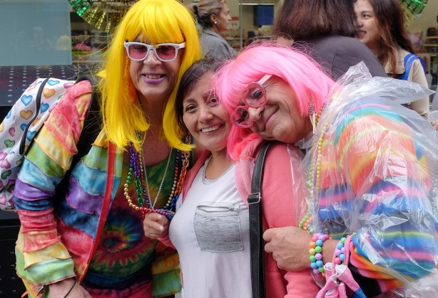 two men in drag pose for a selfie with a woman, one man in bright yellow wig and the other in bright pink with flashy bright clothes to match 