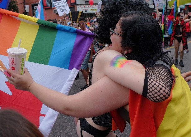 two people hugging at the pride parade, one is a man dressed in a Rocky Horror outfit and the other is a woman with a rainbow flag and a Mcdonalds drink in her hand 