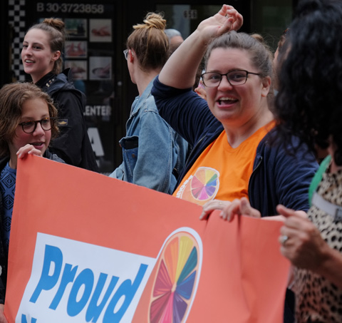 dyke march 2018 - a woman holds an orange Proud NDP sign in the parade, one arm reaching upwards 
