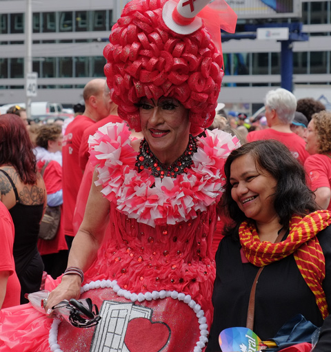 the Queen of Hearts, a drag queen in red with large red plastic wig and a red dress, posses for a pic with a woman 
