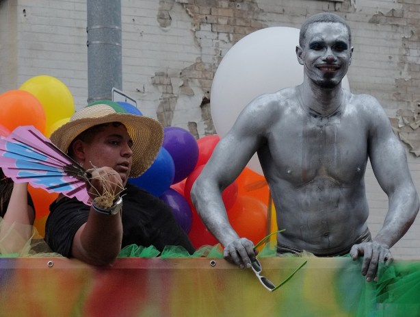 two men on a float at the pride parade, one is covered in silver paint, the other is seated and wearing a large brimmed hat and holding balloons 