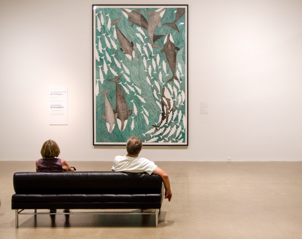 two people sitting on a black sofa, looking at a large painting by Tim Pitsiulak called Swimming with Giants, lots of fish and whales swimming in the water 