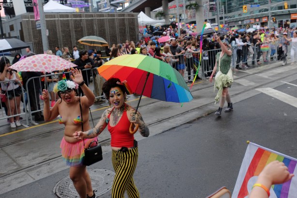 two women walking in the pride parade, crowds on both sides of the street, one is topless with a rainbow frilly skirt and the other has a rainbow umbrella 