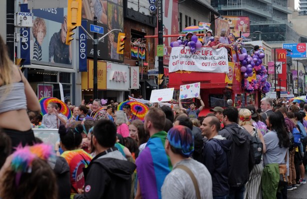the United Church of Canada float in the Pride Parade, people lining the sidewalks on both sides of the street, people walking in front of the float 