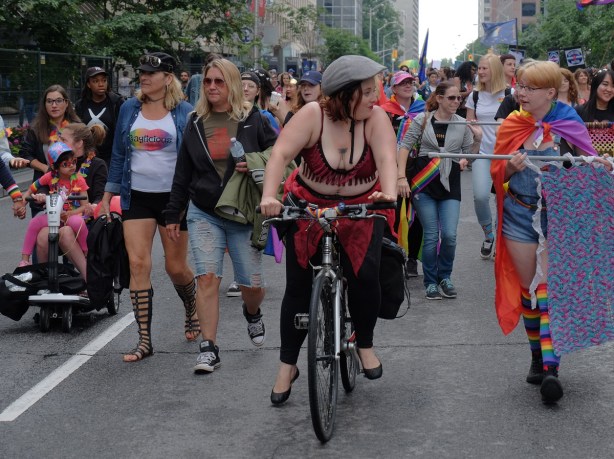 woman on a bike in the dyke march 2018 parade, talking to a young woman who is carrying a banner, other women walking behind, a child on the lap of a woman on a motorized scooter. 