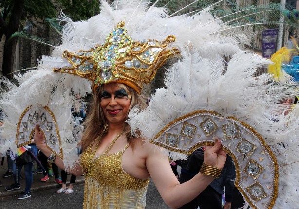 drag queen in elaborate gold costume with white feathers on fans one in each hand) and headdress that also has a large gold glittery star shape front piece 