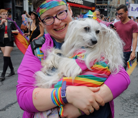 dyke march 2018 - a woman with rainbow headband and pink top is holding a small fluffy white dog with rainbow hat and coat 