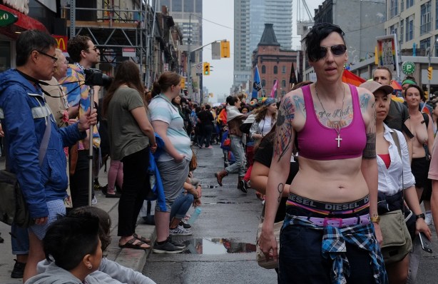 dyke march 2018 - woman in pink bra with many tattoos is walking past a crowd on the sidewalk 