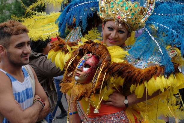 man in drag - blue and yellow feathers, lots of sequins and glitter, carrying a mask 