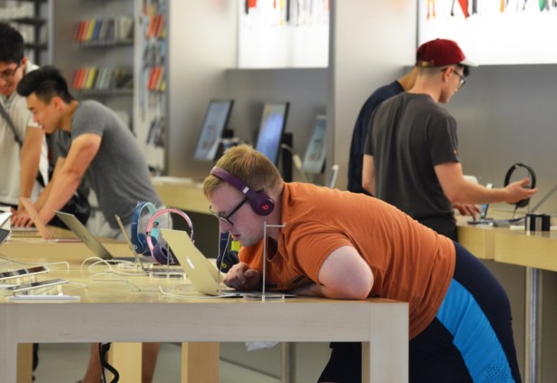 man in an apple store, orange T-shirt, leaning on the counter, with headphones on, looking closely at the screen of a laptop, other men in the store too 