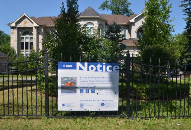 a city of Toronto blue and white notice of development sign is on a black wrought iron fence in front of a large stone faced two storey house with a large front yard, grass and shrubs 