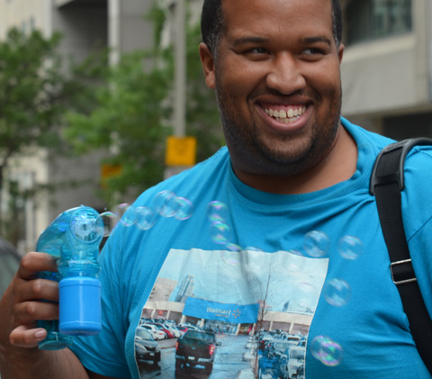 black man with blue t-shirt holds a blue bubble maker and he is making bubbles as he walks down the street