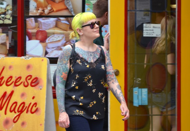 a young woman with bright yellow hair tied back in a short ponytail, tattoo covering most of her arms and upper part of body that is visible , wearing sleeveless black top, walking past Cheese Magic shop in Kensington market, Toronto 