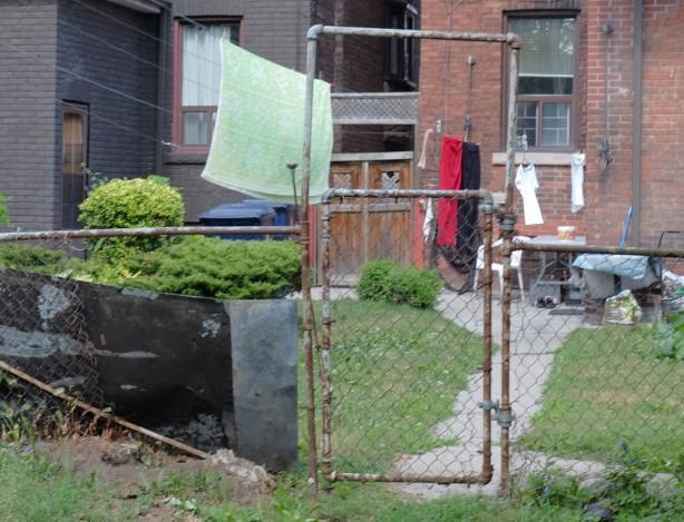 a chainlink fence and gate in a back yard, laundry hanging out to dry in the yard, brick houses, some green grass