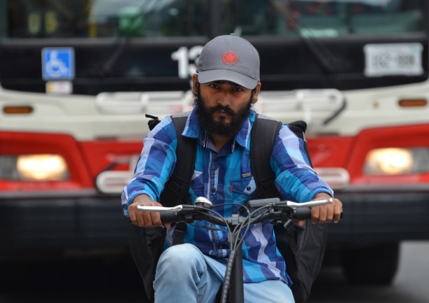 man with beard and moustache, one a bike that is stopped in front of a red and white TTC bus, man wearing a blue baseball cap with red Air Canada maple leaf logo on it 
