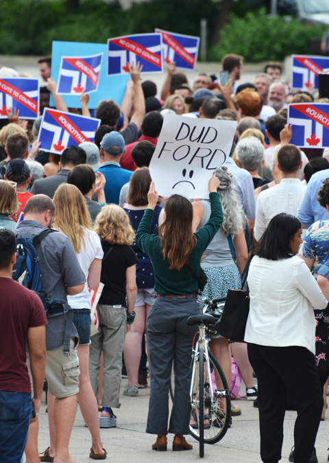 a young woman holds up a sign that says Dud Ford. Words followed by a sad face. Seen from the back. Lots more people in the crowd at the rally 