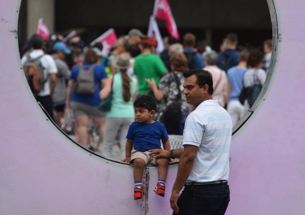 a father and son by the light purple O in the 3D Toronto sign. In the background is a group of people at a protest 