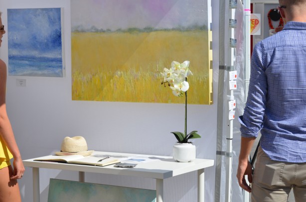 two paintings in a booth at an outdoor art fair, one is yellow, there is a table in front of it, with a flower and and a hat on it, a man walks past 