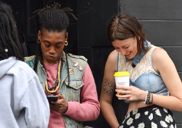 two people, one is a woman holding a coffee cup from Jimmys Coffee, she is laughing. The other is black, hair tied back, pink long sleeved shirt and sleeveless jean jacket, scowling, 