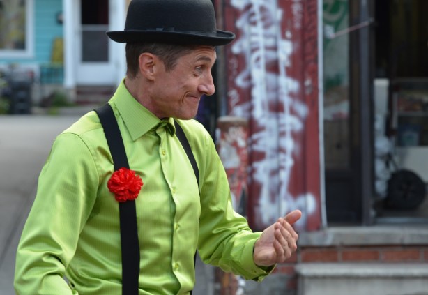 a male mime, black hamburg hat, lime green shirt, black suspenders and red carnation on the suspenders, beckoning to the crowd, outside, street festival, pedestrian sunday at kensington 