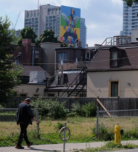 a man walks up Sherbourne street, on the sidewalk, past a vacant lot, in the background is the back of some older brick buildings and beyond that is a tall building with a mural on it, equilibrium by okudart 