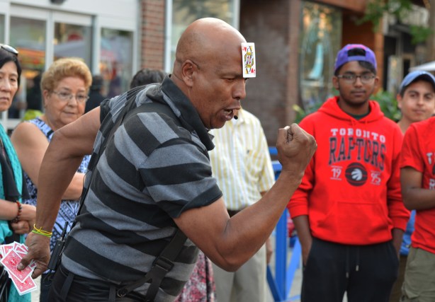 balding black man with the Queen of hearts playing card stuck on his head makes a muscle man stance with fists clenched, he's performing magic tricks while people watch him 