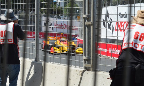 two Honda Indy official photographers get close to the race to take pictures of a yellow race car as it comes around a corner 